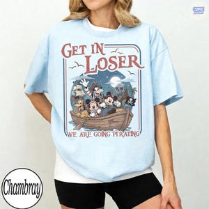 May include: Light blue t-shirt with a pirate-themed graphic featuring cartoon characters in a boat. The design includes the text "Get In Loser We Are Going Pirating" in a red and white font, with a pirate ship and skull.