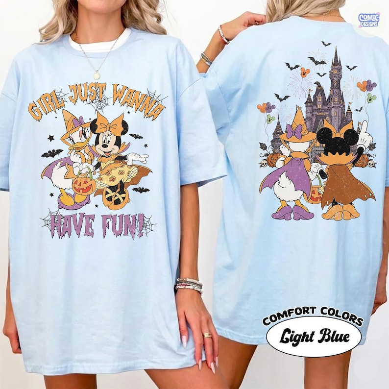 May include: A light blue t-shirt featuring a Halloween-themed graphic. The front displays Daisy Duck and Minnie Mouse in costumes with the text "GIRL JUST WANNA HAVE FUN!" The back shows the characters facing a castle with fireworks and bats.