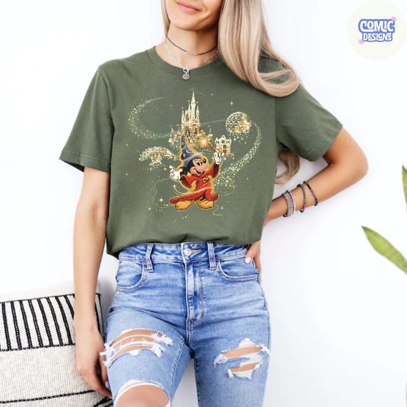 May include: Olive green t-shirt featuring a graphic of Mickey Mouse as a sorcerer with a castle and sparkling accents. The text "COMIC DESIGNS" is in a circle in the upper right corner.