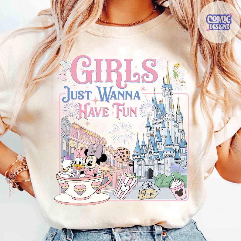 May include: Cream-colored t-shirt featuring a colorful graphic with the text "GIRLS JUST WANNA HAVE FUN." The design includes Minnie and Daisy Duck in a teacup, a castle, fireworks, and other Disney-themed elements.