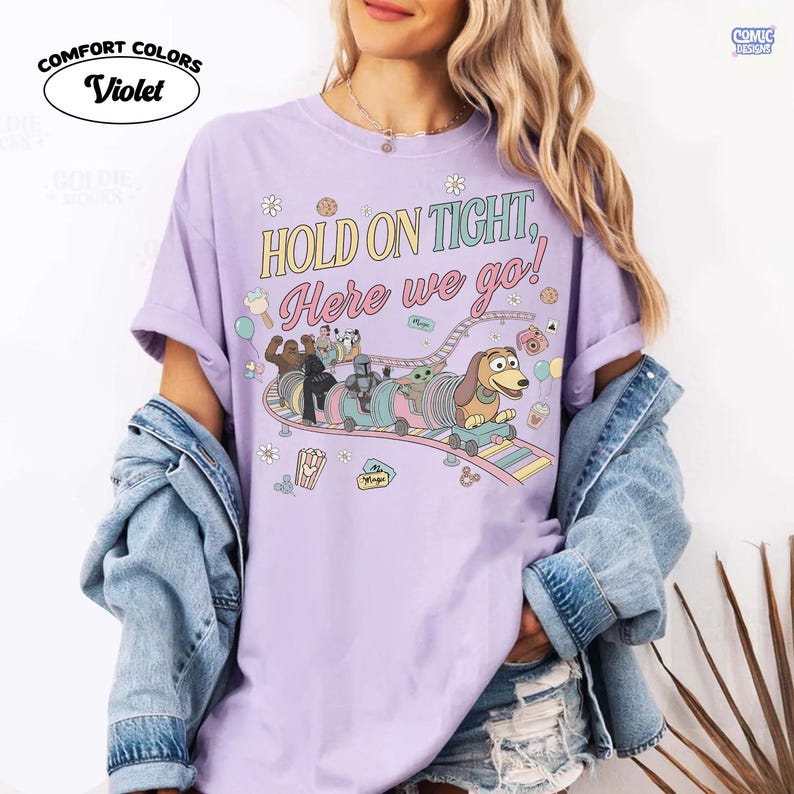 May include: Violet Comfort Colors t-shirt with the text "HOLD ON TIGHT, Here we go!" and a cartoon train design featuring characters. The shirt is light purple and has rolled-up sleeves. The design includes a roller coaster with cartoon characters.