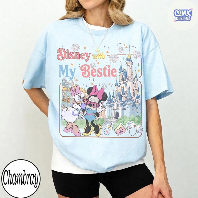 K&ouml;nnte beinhalten: Hellblaues T-Shirt mit einer Grafik von Minnie Mouse und Daisy Duck mit dem Text "Disney with My Bestie". Das Design beinhaltet ein Schloss und andere Disney-Elemente. Das T-Shirt hat kurze &Auml;rmel.