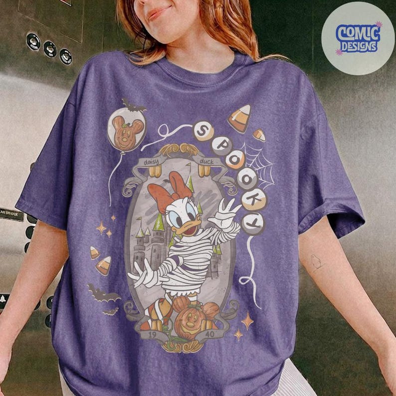 Puede incluir: Una camiseta morada con un gr&aacute;fico de Daisy Duck disfrazada de momia dentro de un marco ornamentado. El dise&ntilde;o incluye el texto SPOOKY, un castillo, dulces de ma&iacute;z, murci&eacute;lagos y un globo con forma de calabaza. Tiene cuello redondo y mangas cortas.