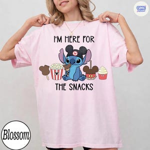 May include: Light pink t-shirt featuring a blue cartoon character wearing a Mickey Mouse hat, surrounded by snack illustrations. The text "I'M HERE FOR THE SNACKS" is printed above and below the character.