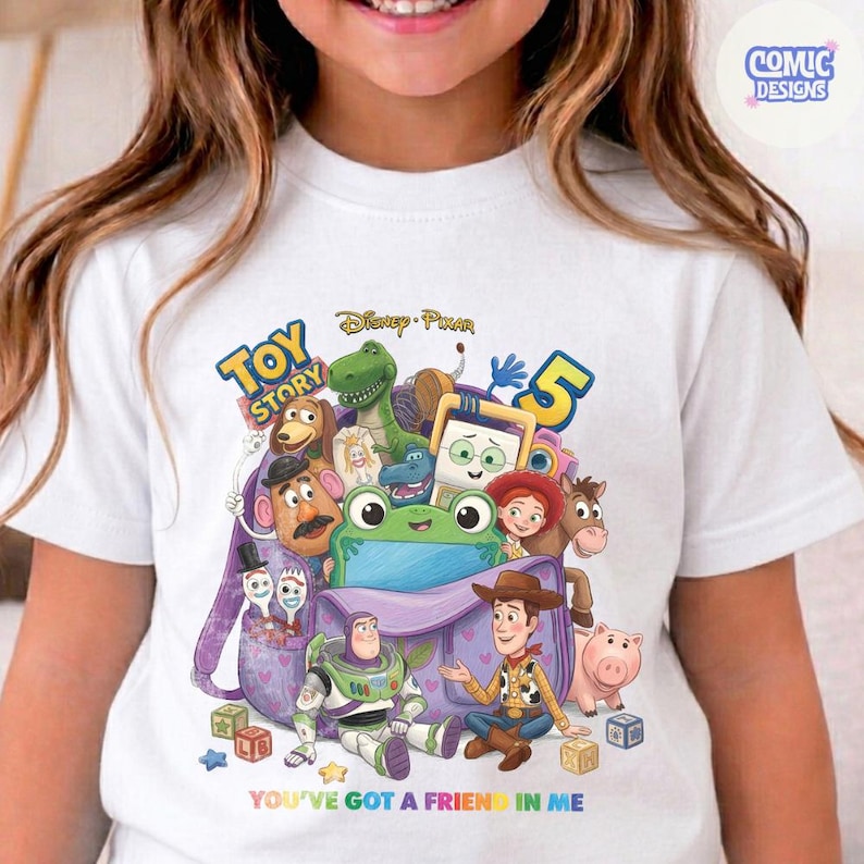 May include: A white t-shirt featuring a colourful graphic of Toy Story 5 characters including Woody, Buzz Lightyear, Rex, and Mr. Potato Head emerging from a purple backpack. The text reads Toy Story 5 and You've Got a Friend in Me.