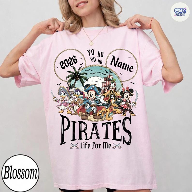 May include: A light pink t-shirt featuring a pirate-themed graphic. The design includes cartoon characters, a pirate ship, and the word "PIRATES" in a bold font. The shirt also has the text "2026" and "Name" in circles.