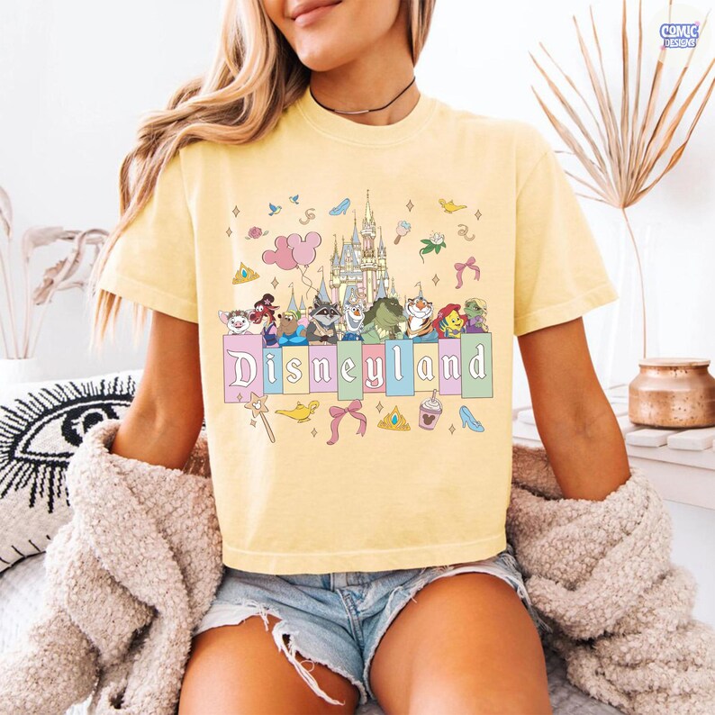 Pu&ograve; includere: T-shirt giallo chiaro con grafica Disneyland. Il design presenta personaggi dei cartoni animati, un castello e la scritta "Disneyland" in colori pastello. Ulteriori grafiche includono una bacchetta magica, fiocchi e una corona.