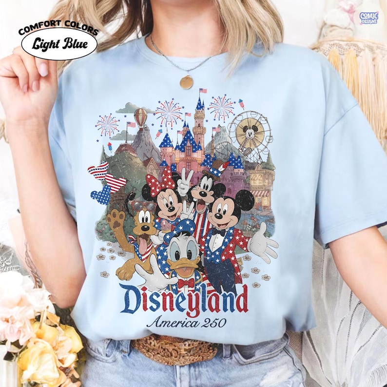 Puede incluir: Una camiseta azul claro con un estampado gr&aacute;fico de Mickey Mouse, Minnie Mouse, Goofy, el Pato Donald y Pluto con vestimenta patri&oacute;tica. El dise&ntilde;o incluye un castillo, fuegos artificiales y el texto Disneyland America 250 en letras rojas y azules.