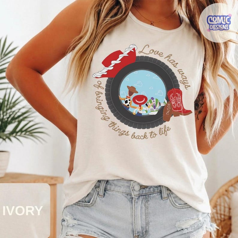 May include: An ivory sleeveless tank top featuring a graphic print of a black tire framing a blue sky scene with cartoon characters. The design includes a red cowboy hat, a red cowboy boot, and the text Love has ways of bringing things back to life.