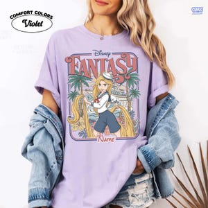 Op de afbeelding: Violet Comfort Colors T-shirt met een Disney Fantasy-ontwerp. De afbeelding toont Rapunzel met lang blond haar, palmbomen en een schip. Het shirt heeft de tekst "Disney Fantasy" en ruimte voor een naam.