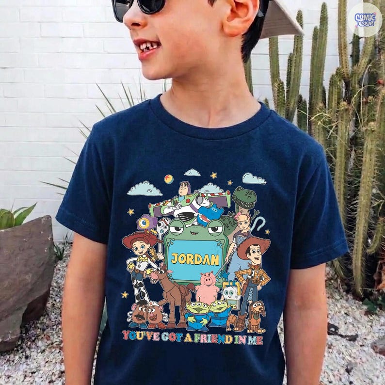 May include: A navy blue t-shirt featuring a colorful graphic print of various animated characters from the Toy Story franchise surrounding a central green frog-shaped frame with the name JORDAN printed in yellow capital letters.