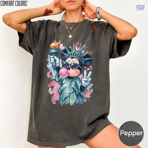 May include: A charcoal gray short-sleeve t-shirt featuring a graphic print of a cartoon mouse dressed as the Statue of Liberty, wearing sunglasses, blowing a pink bubble, holding a torch, and making a peace sign, with abstract pink and blue accents.