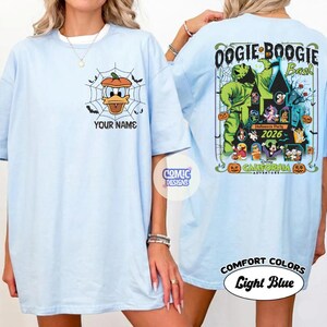 May include: Light blue t-shirts with front and back graphics. The front features Donald Duck in a pumpkin web, with the text "YOUR NAME". The back displays "OOGIE BOOGIE Bash" with Halloween characters and "2026".