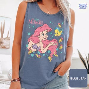 May include: A blue jean colored sleeveless tank top featuring a graphic print of Ariel from The Little Mermaid with pink hair, a purple seashell top, and the yellow fish Flounder surrounded by teal bubbles and the text Disney The Little Mermaid.