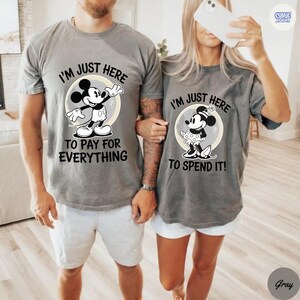May include: Gray t-shirts with Mickey and Minnie Mouse designs. The Mickey shirt says "I'M JUST HERE TO PAY FOR EVERYTHING." The Minnie shirt says "I'M JUST HERE TO SPEND IT!" Both shirts feature a cartoon image of the character inside a circle.