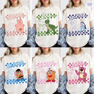 May include: A collection of six off-white t-shirts featuring Toy Story character graphics. Each shirt displays a name in cursive, a checkered border, and a colorful illustration of Hamm, Rex, Bo Peep, Mr. Potato Head, Mrs. Potato Head, or Lotso.
