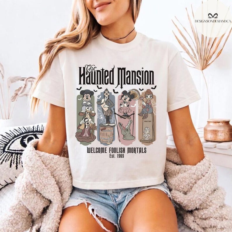 May include: Cream-colored t-shirt featuring "The Haunted Mansion" graphic. The design includes cartoon characters and the text "WELCOME FOOLISH MORTALS Est. 1969". The shirt has short sleeves and a crew neck.