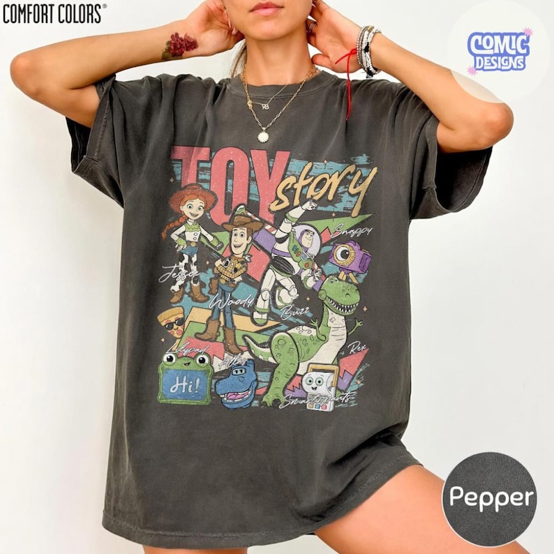 May include: A charcoal gray short-sleeve t-shirt featuring a graphic print of Toy Story characters including Woody, Buzz Lightyear, Jessie, and Rex. The design includes the text Toy Story and character names, with colorful geometric shapes and accents.
