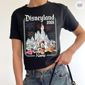 Op de afbeelding: Zwart t-shirt met een kleurrijk Disneyland 2026-ontwerp. Het ontwerp bevat vuurwerk, een kasteel en Disney-personages zoals Mickey en Minnie, Donald en Daisy Duck en Goofy. Het woord "Name" is ook opgenomen.