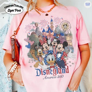 Puede incluir: Una camiseta rosa claro con un estampado gr&aacute;fico de personajes de Disney, incluidos Mickey Mouse, Minnie Mouse, el Pato Donald y Pluto, con ropa patri&oacute;tica roja, blanca y azul. El fondo muestra un castillo, fuegos artificiales y una noria. El texto dice Disneyland America 250.
