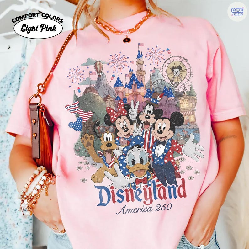 Puede incluir: Una camiseta rosa claro con un estampado gr&aacute;fico de personajes de Disney, incluidos Mickey Mouse, Minnie Mouse, el Pato Donald y Pluto, con ropa patri&oacute;tica roja, blanca y azul. El fondo muestra un castillo, fuegos artificiales y una noria. El texto dice Disneyland America 250.