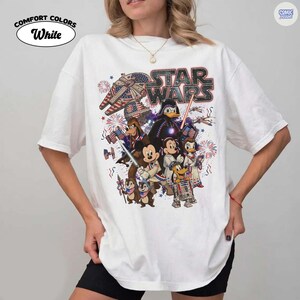 May include: White Comfort Colors t-shirt featuring a Star Wars graphic with Disney characters. The design includes Darth Vader, Mickey Mouse, Donald Duck, and others, with an American flag and fireworks theme.