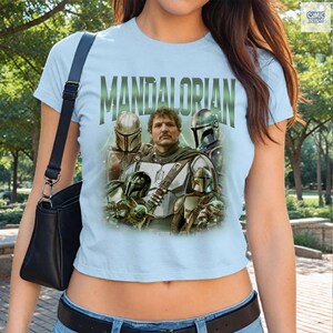 May include: Light blue t-shirt featuring the word "MANDALORIAN" in green block letters. The shirt displays a graphic of characters from the Star Wars series, including the Mandalorian and other figures in helmets.