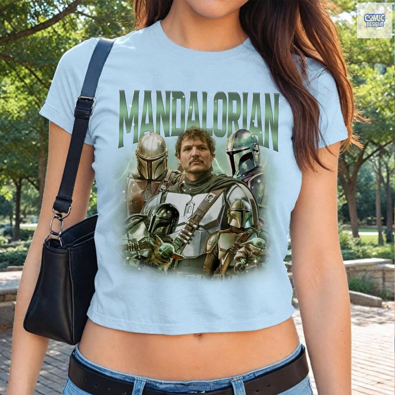 May include: Light blue t-shirt featuring the word "MANDALORIAN" in green block letters. The shirt displays a graphic of characters from the Star Wars series, including the Mandalorian and other figures in helmets.