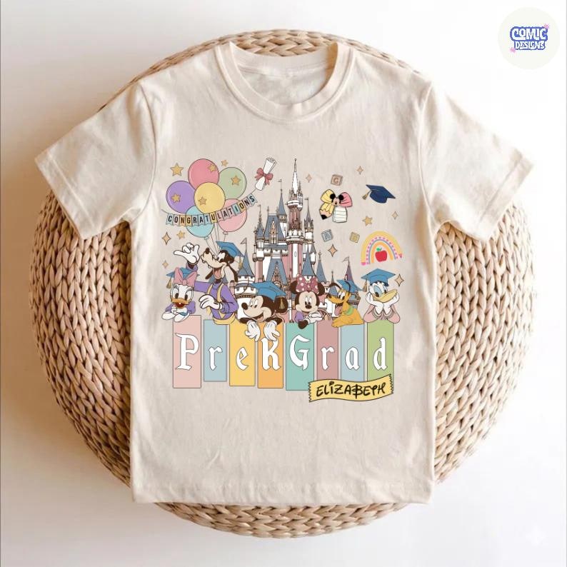 May include: Cream-colored t-shirt featuring a colorful graphic of Disney characters, a castle, balloons, and graduation-themed elements. The text "Prek Grad" and the name "Elizabeth" are also displayed. The shirt is likely for a preschool graduate.