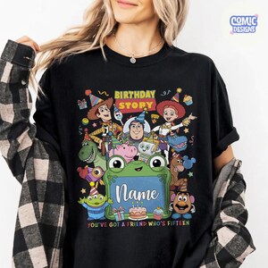 May include: A black t-shirt featuring a colorful graphic of various animated characters wearing party hats. The text reads BIRTHDAY STORY, Name, and YOU'VE GOT A FRIEND WHO'S FIFTEEN. The design includes a green frog-shaped frame and birthday elements.
