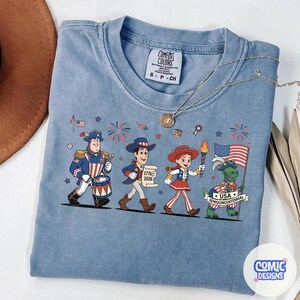 May include: A light blue Comfort Colors t-shirt featuring a patriotic cartoon design. The graphic includes characters in red, white, and blue attire, fireworks, and a waving American flag. The text "USA Semiquincentennial" is also visible.