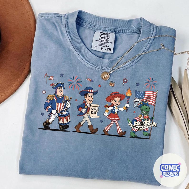 May include: A light blue Comfort Colors t-shirt featuring a patriotic cartoon design. The graphic includes characters in red, white, and blue attire, fireworks, and a waving American flag. The text "USA Semiquincentennial" is also visible.