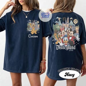 May include: A navy blue t-shirt featuring a small graphic on the front chest of Pluto in front of a castle with the text Custom. The back displays a large graphic of Mickey Mouse, Minnie Mouse, Donald Duck, and Pluto at Disneyland with the text Disneyland Est. 1955.