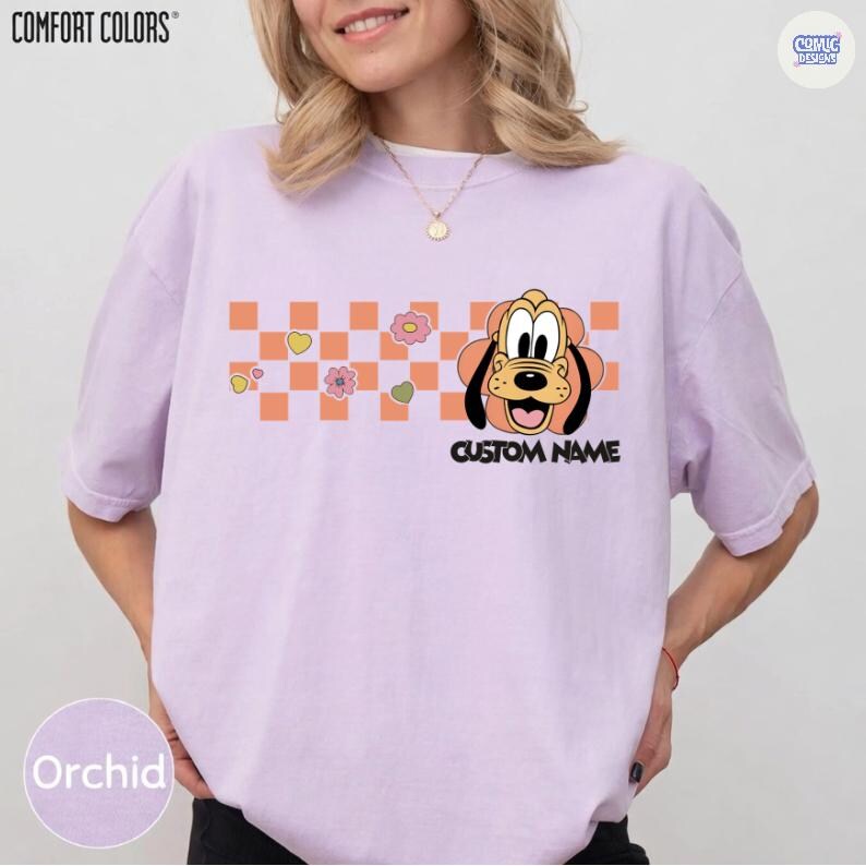May include: Orchid Comfort Colors t-shirt featuring a cartoon dog design with a custom name option. The design includes a checkerboard pattern with flowers and hearts. The shirt is a light purple color.