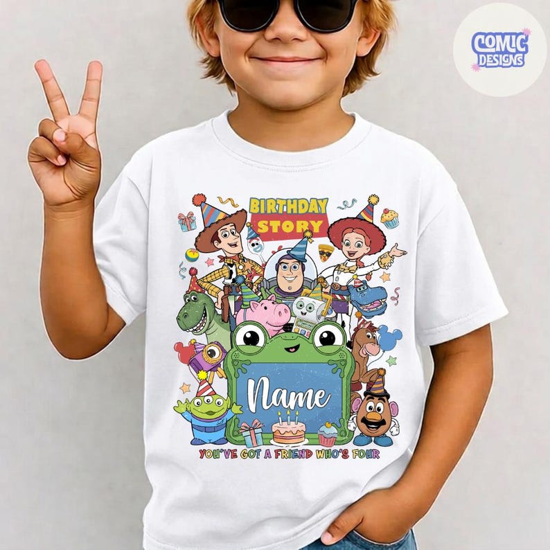 May include: A white t-shirt featuring a colorful graphic of various animated toy characters wearing party hats. The design includes the text "BIRTHDAY STORY", "Name", and "YOU'VE GOT A FRIEND WHO'S FOUR" centered above a green frog-shaped frame.