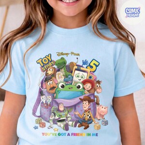 May include: A light blue t-shirt featuring a graphic of Toy Story characters emerging from a purple backpack. The design includes Buzz Lightyear, Woody, Rex, and other figures, with the text Toy Story 5 and You've Got a Friend in Me.