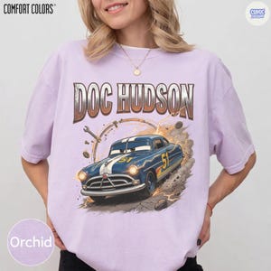 Pode incluir: Camiseta cor de orqu&iacute;dea com um gr&aacute;fico do carro de Doc Hudson do filme Carros. O design inclui o texto "DOC HUDSON" acima do carro, com o n&uacute;mero "51" do carro vis&iacute;vel. A camisa tem os logotipos "COMFORT COLORS" e "COMIC DESIGN".