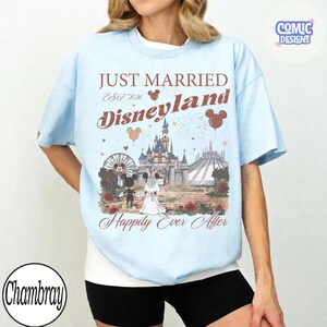 May include: Light blue t-shirt with a Disneyland-themed graphic. The design includes the text "Just Married Disneyland Happily Ever After" with Mickey and Minnie Mouse, a castle, and fireworks. The shirt has a crew neck and short sleeves.