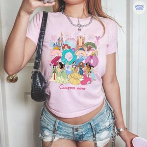 May include: A light pink short-sleeve t-shirt featuring a colorful graphic of various animated princesses, a castle, and the year 2024. The design includes the text Custom Name below the characters. The shirt has a crew neckline and a fitted cut.