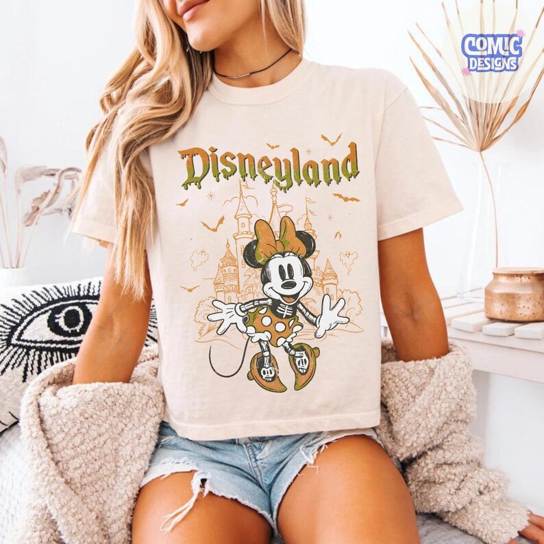 Może przedstawiać: Kremowy t-shirt z grafiką szkieletu Myszki Minnie w sp&oacute;dniczce w kropki i kokardce. Nad postacią znajduje się napis Disneyland zieloną, ściekającą czcionką, a w tle zarys zamku i małe nietoperze.
