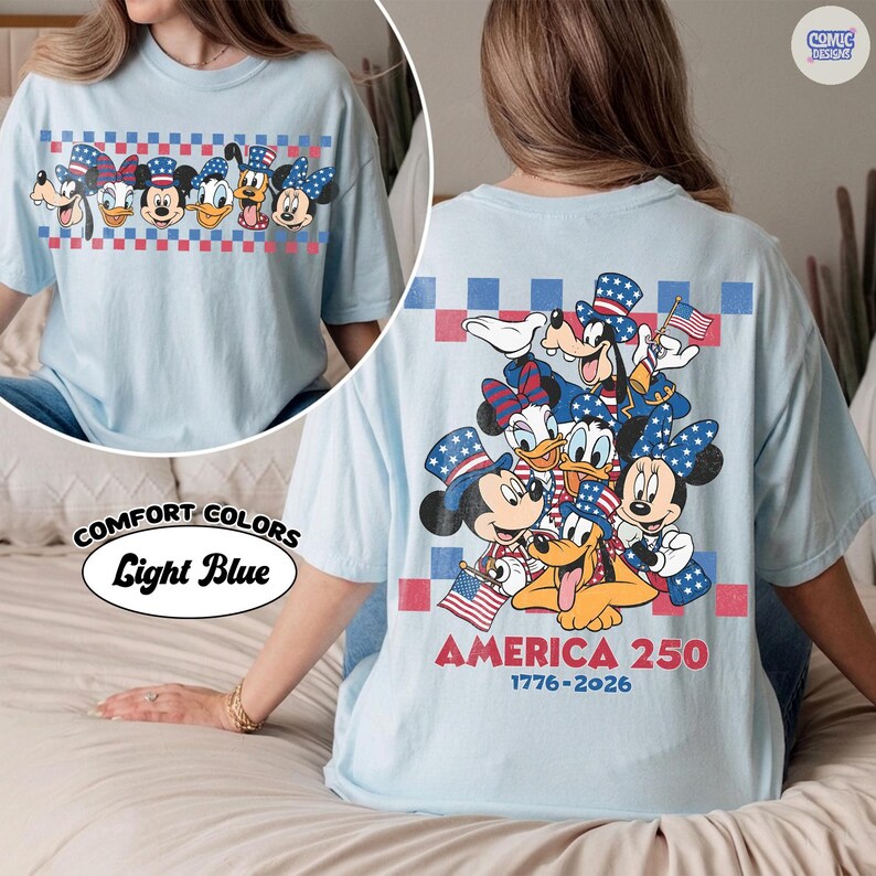 May include: Light blue t-shirt featuring cartoon characters in patriotic attire. The front displays a checkered pattern with characters. The back has a group of characters with flags and the text "AMERICA 250 1776-2026". The shirt is labeled "Comfort Colors Light Blue".