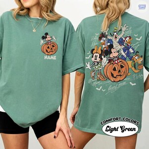 May include: A sage green t-shirt featuring a small graphic of Mickey Mouse sitting on a pumpkin on the front left chest. The back displays a large illustration of various Disney characters in Halloween costumes around a pumpkin with the text NAME.
