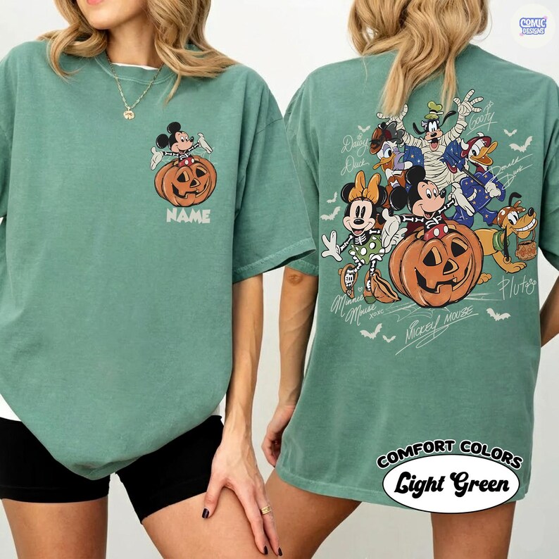 May include: A sage green t-shirt featuring a small graphic of Mickey Mouse sitting on a pumpkin on the front left chest. The back displays a large illustration of various Disney characters in Halloween costumes around a pumpkin with the text NAME.