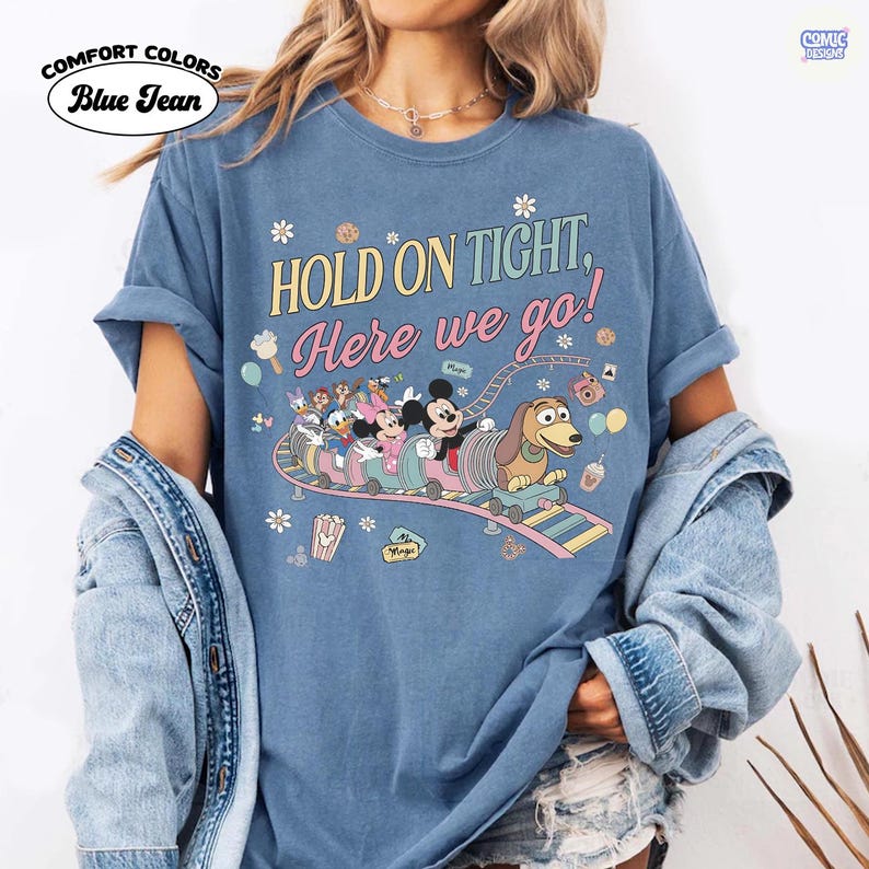 May include: Blue, Comfort Colors t-shirt with the text "HOLD ON TIGHT, Here we go!" and a cartoon roller coaster design featuring Disney characters. The shirt has rolled-up sleeves and a relaxed fit.