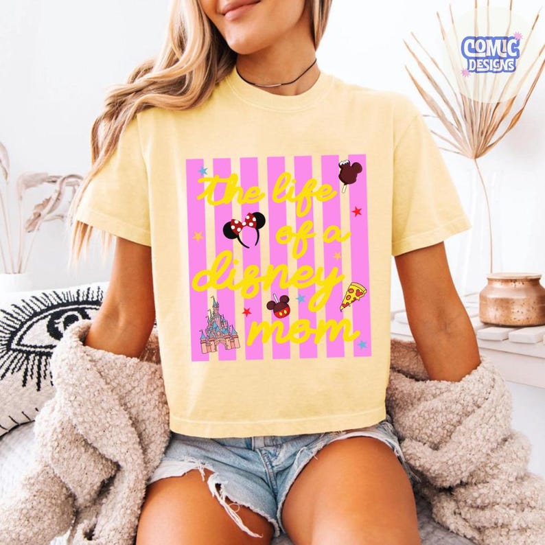 Puede incluir: Camiseta amarilla clara con el texto "The life of a Disney mom" en letras amarillas. El dise&ntilde;o incluye rayas rosas, estrellas e iconos de Disney como orejas de Mickey, un castillo y una porci&oacute;n de pizza.