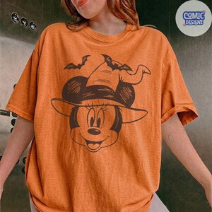 May include: Orange t-shirt featuring a cartoon Minnie Mouse wearing a witch hat and two bats. The graphic is in dark brown. The shirt has short sleeves and a round neckline. The text "COMIC DESIGNS" is in a circle in the upper right corner.