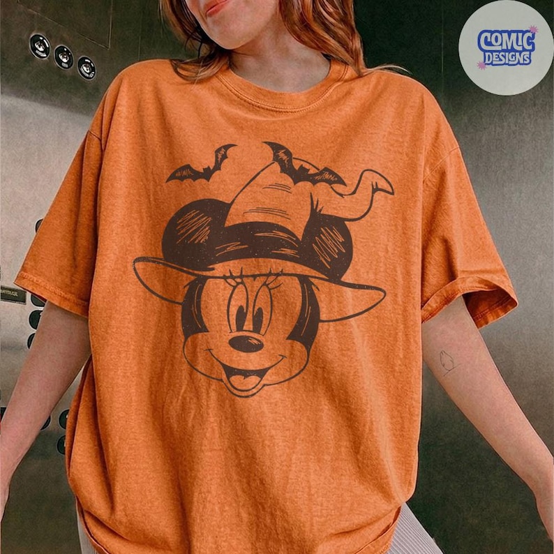 May include: Orange t-shirt featuring a cartoon Minnie Mouse wearing a witch hat and two bats. The graphic is in dark brown. The shirt has short sleeves and a round neckline. The text "COMIC DESIGNS" is in a circle in the upper right corner.