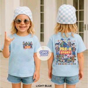 May include: Light blue t-shirt with a cartoon graphic on the front and back. The front graphic says "NAME" and features cartoon characters in graduation attire. The back graphic says "PRE-K STORY TO KINDERGARTEN AND BEYOND".