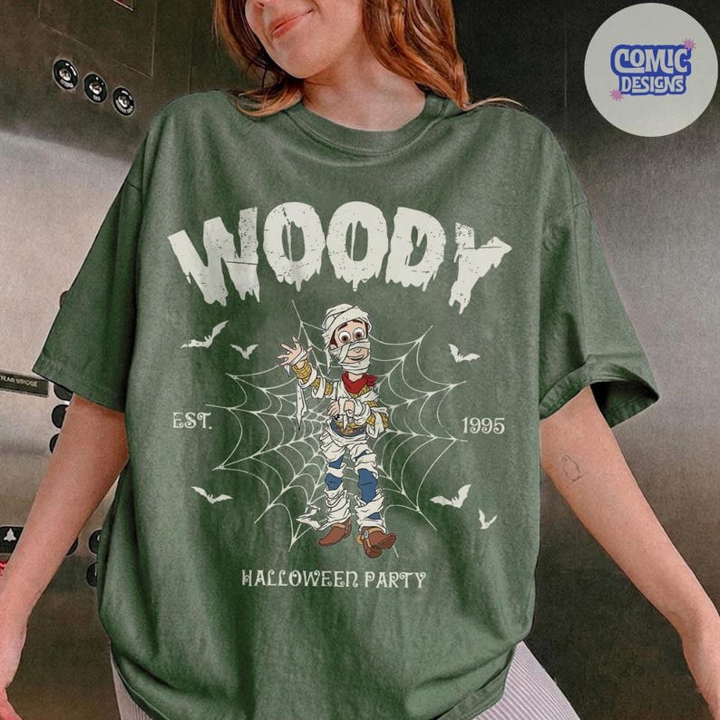 May include: An olive green short-sleeve t-shirt featuring a graphic of a character wrapped in bandages like a mummy standing in a spiderweb. The text reads WOODY, EST. 1995, and HALLOWEEN PARTY, accompanied by small white bat illustrations.