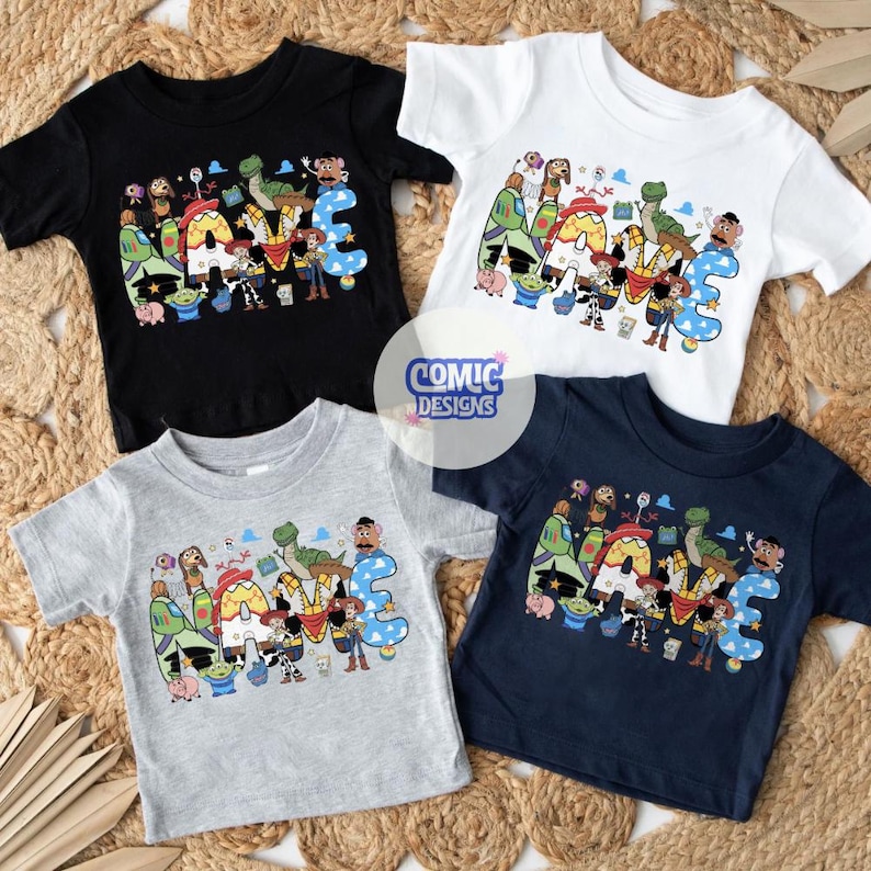 May include: Four short-sleeved t-shirts in black, white, grey, and navy blue. Each shirt features a graphic print of the word NAME with colourful cartoon characters from Toy Story integrated into the letters. A circular logo reads COMIC DESIGNS.